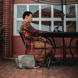 White Duck Outdoors Filios Messenger Bag Olive on the floor next to a man outside a cafe