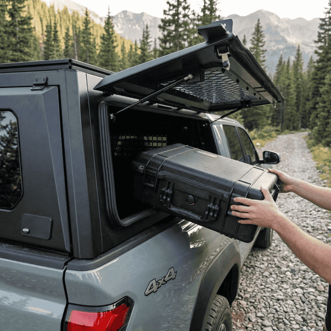 Overland Vehicle Systems Expedition Truck Cap with Sliding Windows for 2016 to 2023 Toyota Tacoma 5 Ft. Bed parked with a man storing a box through the window