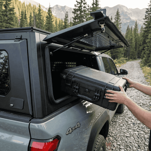 Overland Vehicle Systems Expedition Truck Cap with Sliding Windows for 2016 to 2023 Toyota Tacoma 5 Ft. Bed parked with a man storing a box through the window