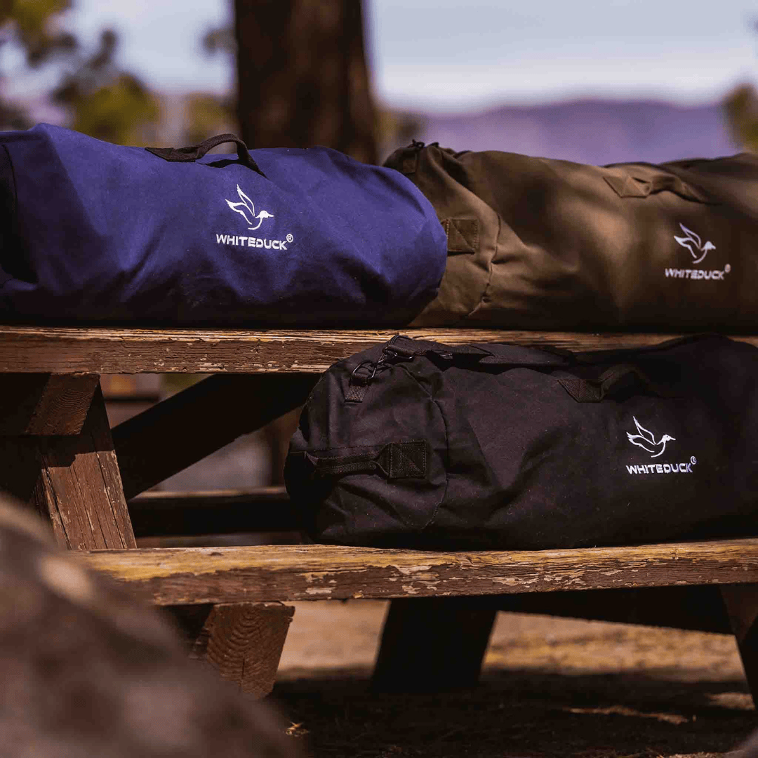 White Duck Outdoors Hoplite Canvas Duffel Bag blue black and olive on bench